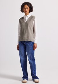 Kvinna som står klädd i en vit skjorta med knappar, grå ärmlös tröja, blå jeans och vita sneakers mot en enfärgad bakgrund.