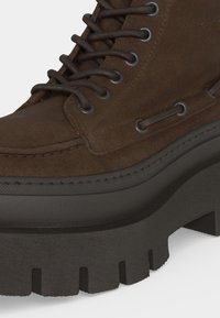 Botte en daim marron avec une semelle épaisse noire à crampons, un bout rond et des œillets métalliques avec des lacets marron assortis en gros plan.
