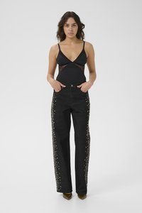 Mujer de pie mirando hacia adelante, lleva una blusa sin mangas negra con lados transparentes y jeans negros adornados con tachuelas plateadas a lo largo de las piernas.
