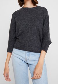 Mörkgrå stickad tröja med rund halsringning och trekvartsärmar, i kombination med ljusblå jeans. Mjuk textur och avslappnad passform.