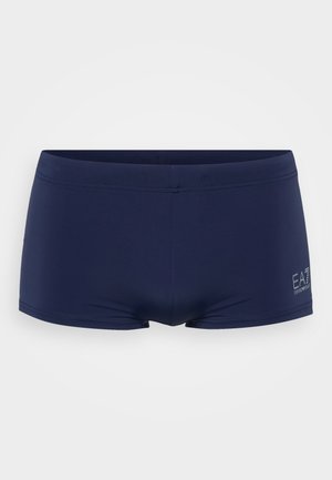 Bañador de color azul marino hecho de tela elástica, con una cintura baja y un pequeño logo en el lateral. Textura suave y diseño ajustado.