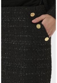 Černá strukturovaná látka s kovovými nitěmi, se třemi zlatými knoflíkovými ozdobami poblíž kapsy. Detailní záběr na střih a materiál sukně.