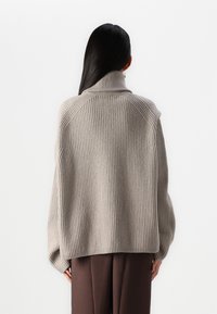Maglione beige a coste con colletto alto e spalle cadute; texture morbida; vestibilità comoda; indossato sopra pantaloni marroni.