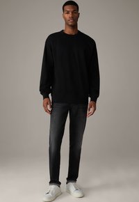 Schwarzer Langarm-Cotton-Pullover, lockere Passform, kombiniert mit dunkelgrauen Jeans und weißen Sneakers, vor einem neutralen Hintergrund präsentiert.