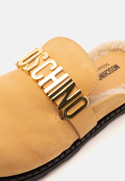 Chaussure à enfiler couleur sable avec semelle noire, lettres dorées "MOSCHINO" sur le dessus et doublure intérieure en shearling beige doux.