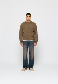 Hnedý sveter s rebrovaným výstrihom, skombinovaný s modrými rovnými džínsami. Model má na sebe svetlú obuv. Minimálny pozadí.