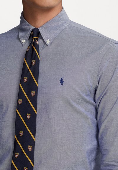 Svetložna modra srajca z ovratnikom, ki ima vezen temno modri logotip. Zasnovana je z mornarsko kravato z rumenimi diagonalkami in vzorci ščita.