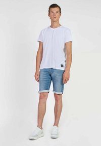 T-shirt en coton blanc, short en denim bleu avec ourlet roulé, baskets blanches avec accents gris, se tenant sur un fond uni.