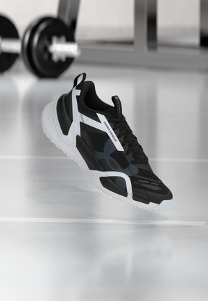 Schwarz-weißes Under Armour Sportschuh mit Schnürsenkeln schwebt über einem reflektierenden Fitnessraum-Boden, eine Stange mit Gewichten im Hintergrund unscharf.