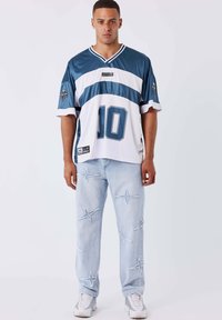 Giovane uomo che indossa una maglietta sportiva blu e bianca con il numero 10, jeans azzurri chiari con toppe a forma di stella e sneaker bianche, in piedi su uno sfondo bianco.