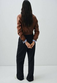 Veste aviateur en cuir marron avec des poignets côtelés, associée à un pantalon large à rayures noires. Mains jointes derrière le dos.