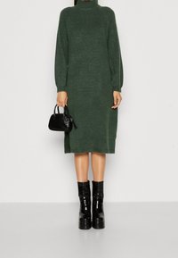 Persona che indossa un lungo vestito a collo alto di colore verde scuro, tiene una piccola borsa nera e indossa stivaletti neri a piattaforma lucidi.