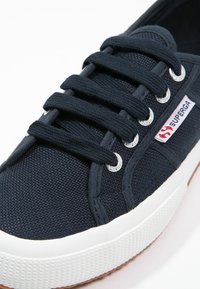 Marinblå canvas sneakers med en texturerad ovansida, vit gummisula och silverfärgade öglor. Har platta snören och en röd/vit logomärke.