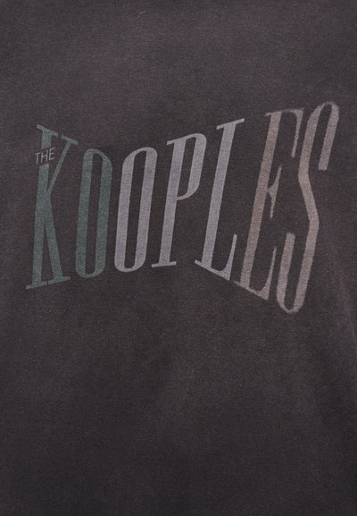 Σκούρο ύφασμα με μεγάλα, καμπύλα γράμματα που γράφουν "THE KOOPLES" με κεφαλαία γράμματα στο κέντρο.