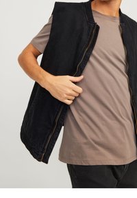 Czarna, bezrękawnik z zamkiem z przodu i prążkowanym kołnierzem, założony na jasnobrązowy t-shirt, o gładkiej teksturze i minimalistycznym designie.