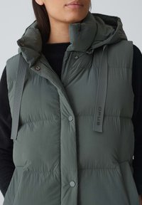 Khakifarbene Steppweste mit hohem Kragen und Kapuze. Verfügt über Knopfverschlüsse, vertikale Nähte und verstellbare Riemen mit Branding an der Seite.