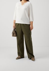 Biała bluzka z wzorem w dziurki, z rękawami o długości trzy czwarte, w zestawieniu z luźnymi, oliwkowymi spodniami cargo oraz beżowymi butami typu slip-on.