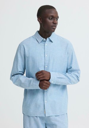 Mężczyzna w jasnoniebieskiej koszuli z długim rękawem zapinanej na guziki i dopasowanych spodniach, patrzący w bok z rękami złożonymi z przodu.