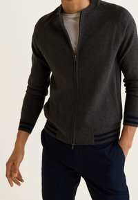 Homme portant un pull zippé gris foncé avec des poignets et un ourlet rayés, un sous-pull blanc et un pantalon bleu marine foncé, debout avec une main dans la poche.