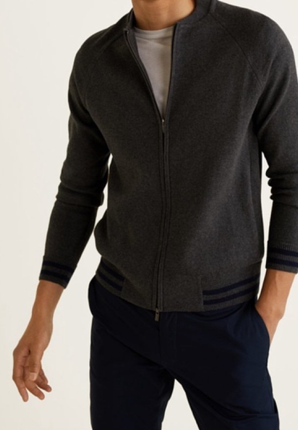 Homme portant un pull zippé gris foncé avec des poignets et un ourlet rayés, un sous-pull blanc et un pantalon bleu marine foncé, debout avec une main dans la poche.