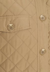 Beige gewatteerde jas met een borstzak met knoopsluiting, twee grote knopen aan de voorkant, een gladde textuur en een diagonale stikpatroon.