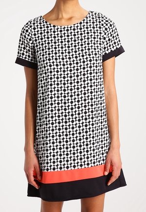 Kvinde iført en kortærmet knælængde kjole med sort-hvidt cirkelmønster, koralfarvet stribe og sort kant.