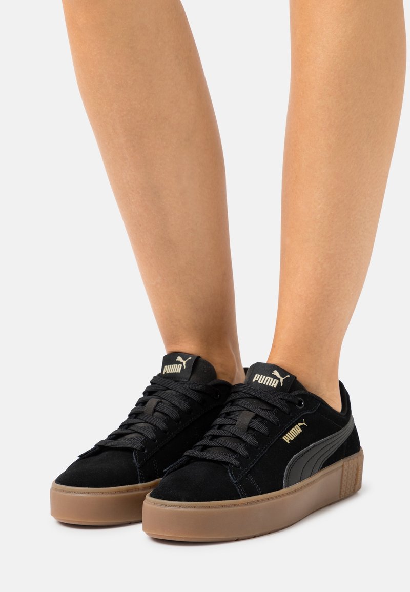 Baskets noires Puma avec semelles en gomme portées pieds nus, montrant les lacets et le logo Puma sur la languette et sur le côté.