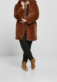 Brun faux fur frakke med hætte, der har en blød tekstur og åben frontdesign. Sammen med sorte bukser og brune ankelstøvler.