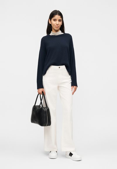 Γυναίκα που στέκεται κρατώντας μαύρη τσάντα, φοράει μπλε πουλόβερ πάνω από ριγέ ζιβάγκο, λευκό παντελόνι και λευκά αθλητικά παπούτσια με μαύρες λεπτομέρειες.