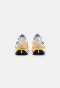Baskets sportives avec un dessus léger de couleur blanc cassé, un col bleu, des détails noirs, et une semelle orange et jaune avec une texture de type crampons.