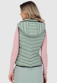 Gilet imbottito verde chiaro con cappuccio, caratterizzato da un motivo a zig zag, orlo elastico e rifiniture in contrasto. Indossato sopra un top a maniche lunghe rosa.