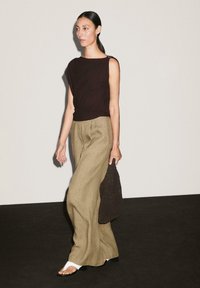 Haut sans manches marron, pantalon large en lin beige, et sandales blanches à enfiler. Portant un sac en cuir texturé marron, posé sur un fond clair.