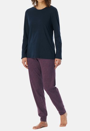 Person wearing a navy long-sleeve shirt and purple patterned pants, standing barefoot on a light background.