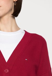 Kvinna i vit skjorta under en röd kofta med knappar och en liten rektangulär logotyp på bröstet, mot en enfärgad bakgrund.