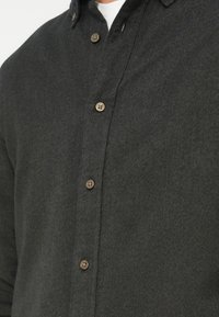 Mørkegrønn skjorte med knapper og strukturert stoff, med brune knapper og krage, delvis synlig i den øvre delen av bildet.