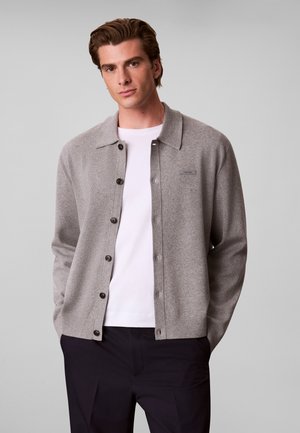 Jeune homme aux cheveux bruns portant un cardigan gris boutonné par-dessus un t-shirt blanc et un pantalon noir sur un fond gris uni.