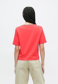 Hosszú fonott hajú nő vörös rövid ujjú ingben és világos bézs nadrágban, háttal áll a kamerának fehér háttér előtt.