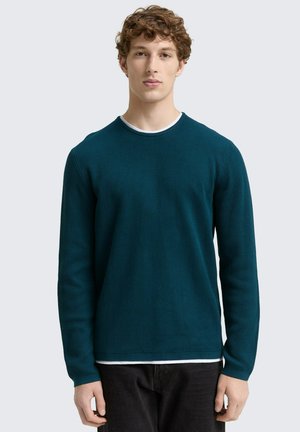 Türkis gestreiftes Langarmshirt mit Rundhalsausschnitt und kontrastierendem weißen Innenkragen. Getragen mit dunklen Hosen, die ein tailliertes Design präsentieren.