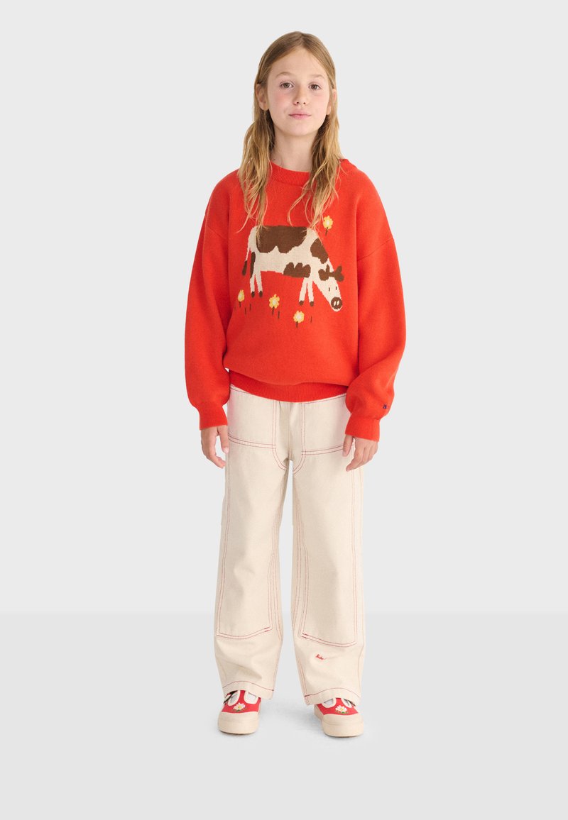 Jeune personne portant un pull rouge avec un motif de vache et de fleurs, un pantalon ample beige clair avec une couture rouge, et des chaussures rouges et blanches.