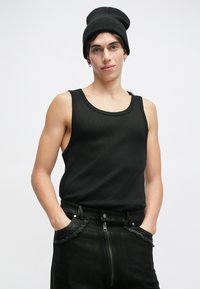 Crna majica bez rukava s rebrastom teksturom, okruglim izrezom, nosi se uz tamne traperice s patentnim zatvaračem i isfrendanim rubovima. Uz to se nosi crna kapa.