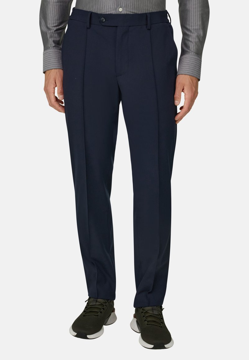 Homme portant un pantalon bleu marine sur mesure, une chemise grise rayée à manches longues et des chaussures de sport vert foncé, debout devant un fond uni.