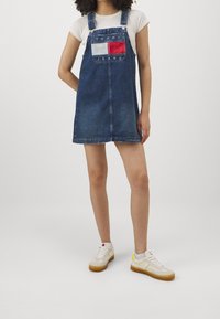 Džínové šaty s laclem v tmavě modré barvě, s přední kapsou s červenými a bílými detaily. V kombinaci s bílými teniskami se žlutými prvky.