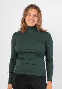 Maglietta a maniche lunghe verde scuro con collo alto realizzata in un tessuto liscio e elastico, con una vestibilità aderente e dettagli di cucitura minimi. Abbinata a leggings neri lucidi.