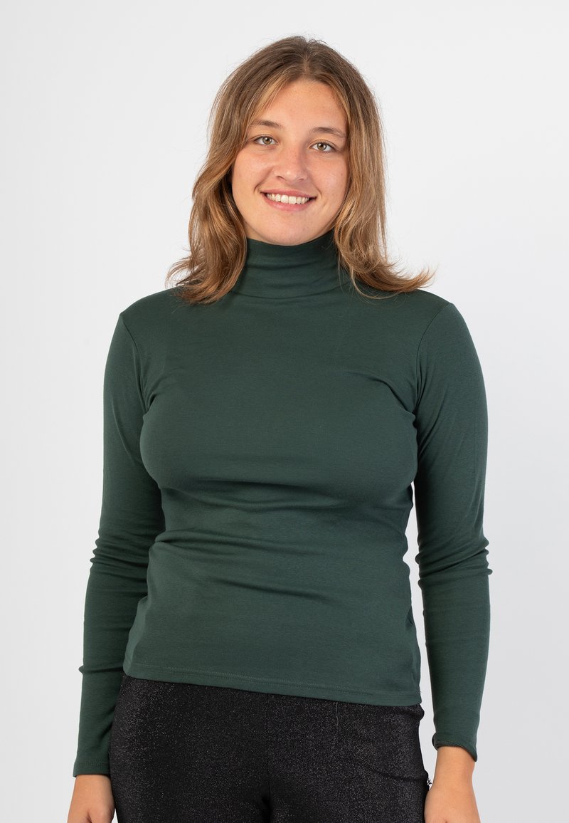 Maglietta a maniche lunghe verde scuro con collo alto realizzata in un tessuto liscio e elastico, con una vestibilità aderente e dettagli di cucitura minimi. Abbinata a leggings neri lucidi.