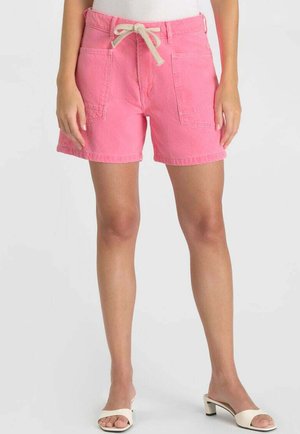 Woman wearing pink drawstring shorts with large front pockets and white open-toe block heel sandals, against a plain background.