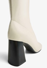 Botine crem cu toc, având un design curbat, toc bloc texturat negru și cusături vizibile de-a lungul cusăturii. Material moale și neted.