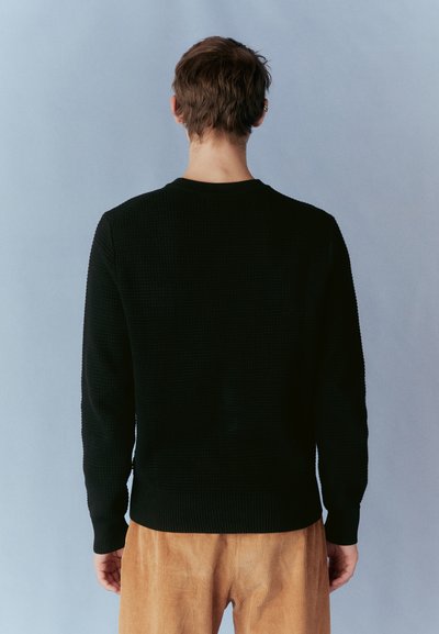 Pulover negru țesut, cu manșete și tiv cu lână, având un model texturat. Modelul poartă pantaloni din catifea, fiind prezentat din spate, pe un fundal albastru.