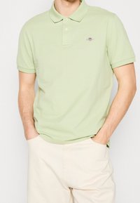 Homme portant un polo à manches courtes vert clair avec un petit logo brodé sur la poitrine, accompagné d'un pantalon beige clair, les mains dans les poches.