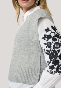Gray knitted vest with a ribbed hem, featuring decorative buttons on the side, worn over a white shirt with black floral embroidery.