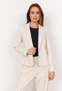 Blazer beige con vestibilità su misura, revers a punta e due bottoni. Indossato sopra un top scuro a coste, abbinato a pantaloni beige coordinati.